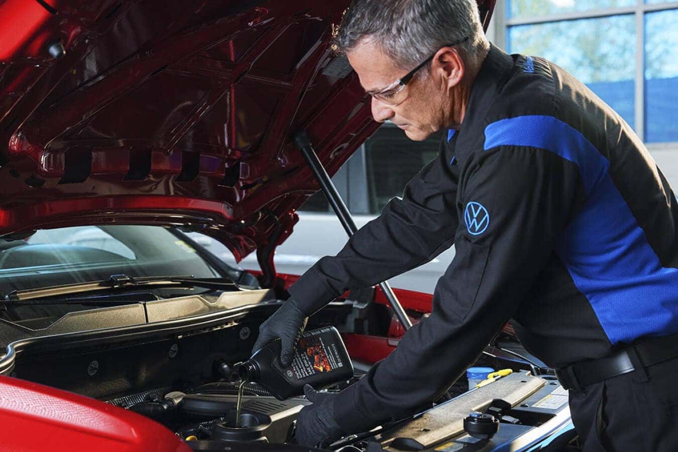 A Volkswagen Service Technician pouring oil into the engine of a VW vehicle with the hood up.