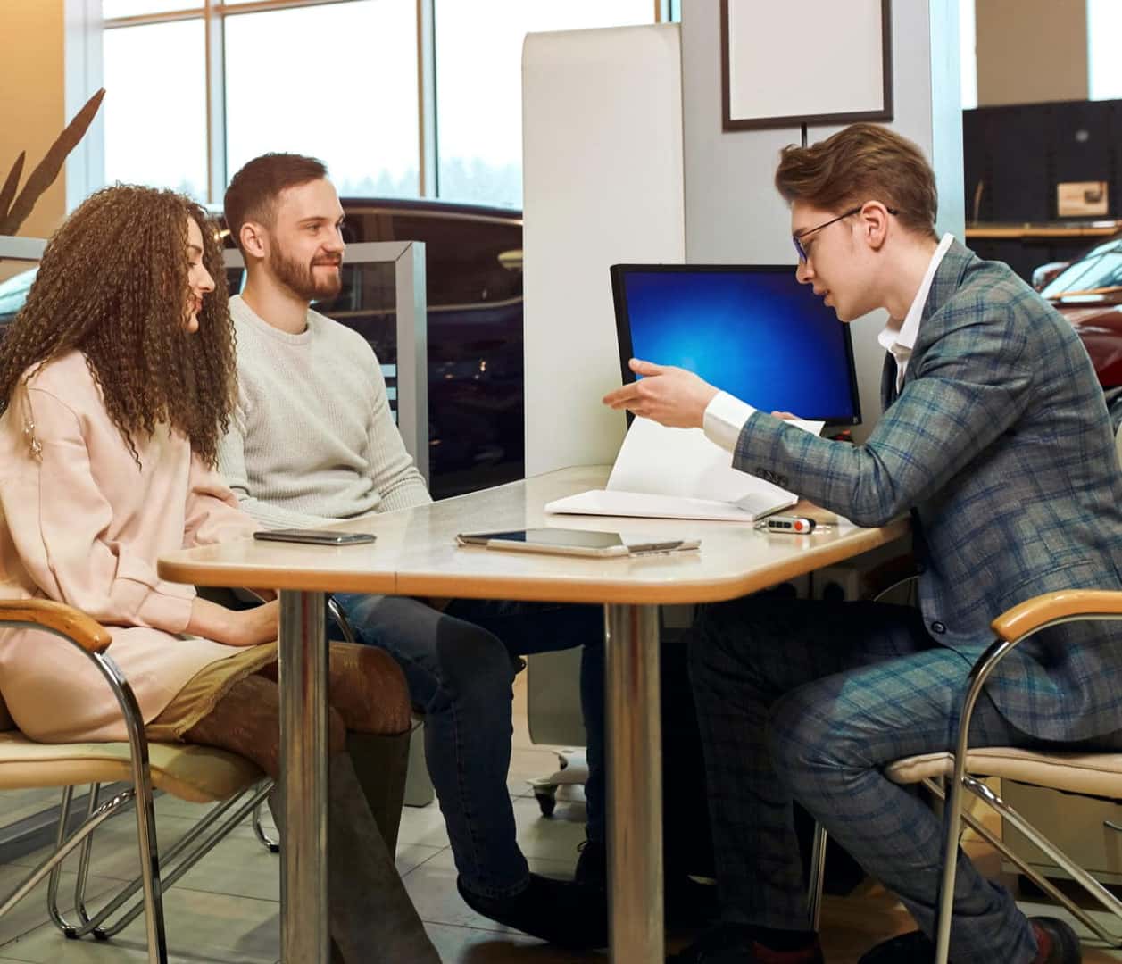 Car salesperson discussing paperwork with a couple seated at a desk inside a dealership.