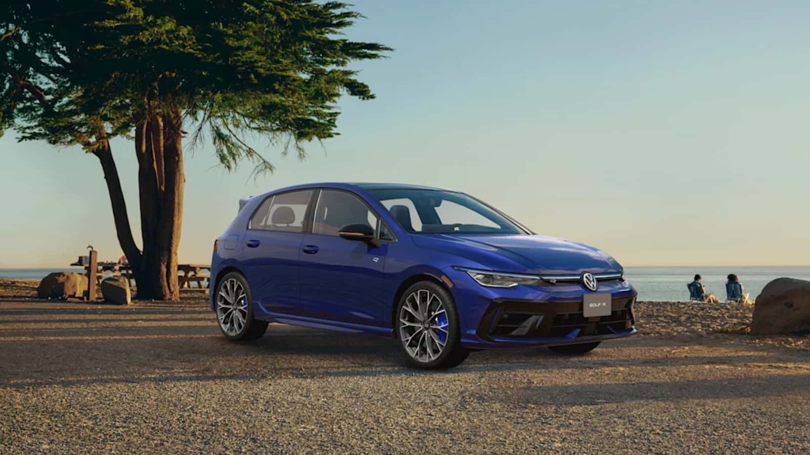 2026 Volkswagen Golf R performance AWD hatchback parked along a scenic coastal beach road overlooking the ocean with couple relaxing nearby, available at Midlands Volkswagen in Columbia, SC