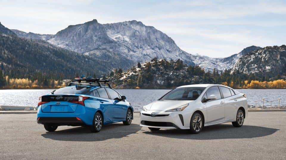 A white 2020 Prius and a blue 2020 Prisu against a mountain and lake.