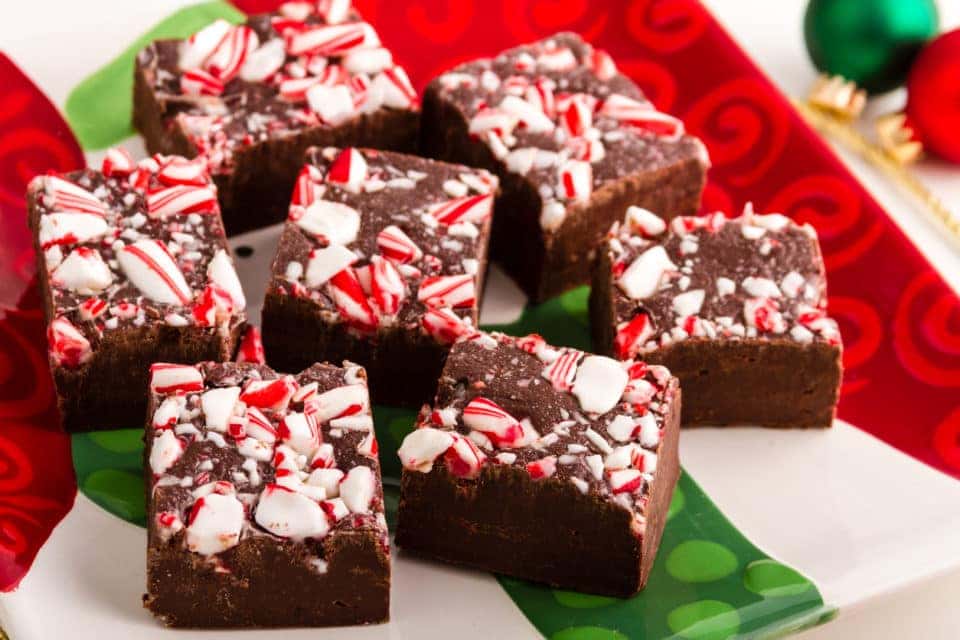 A pile of chocolate fudge with crushed candy cane on top. The fudge is sitting on a white, red, and green-striped holiday plate.