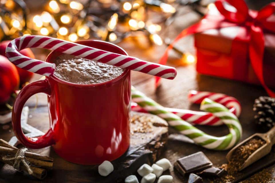 Red mug filled with homemade hot chocolate shot on rustic Christmas table. A red and white candy cane is on the hot chocolate mug and two others candy canes are behind the mug. String Christmas lights and a red gift are out of focus on background. Predominant color is red. DSRL studio photo taken with Canon EOS 5D Mk II and Canon EF 100mm f/2.8L Macro IS USM.