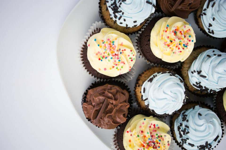 nine chocolate cupcakes on a white plate on a white background. One cupcake has chocolate frosting, two have yellow frosting, and four have blue frosting.