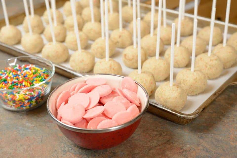 A bowl with pink candy melts, a bowl with rainbow Jimmy sprinkles, and a half sheet tray with un-iced vanilla cake pops.