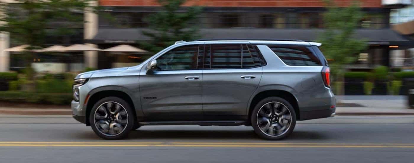 A silver 2025 Chevy Tahoe is shown driving on a city street to visit a Chevy dealer.