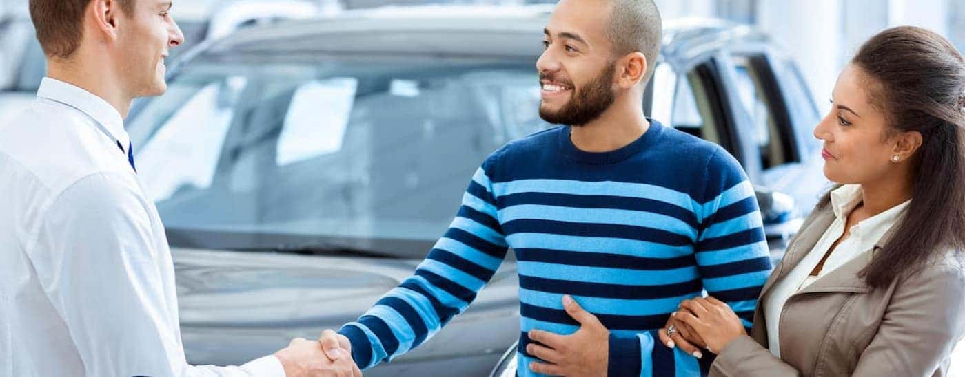 A salesman is shaking hands with a husband who is with his wife in front of used cars.