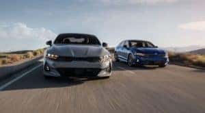 A gray and a blue 2021 Kia K5 are on a desert highway and shown from the front.