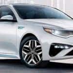 A white 2020 Kia Optima, one of the hybrid Kia car models, is parked in front a white wall with art on it.