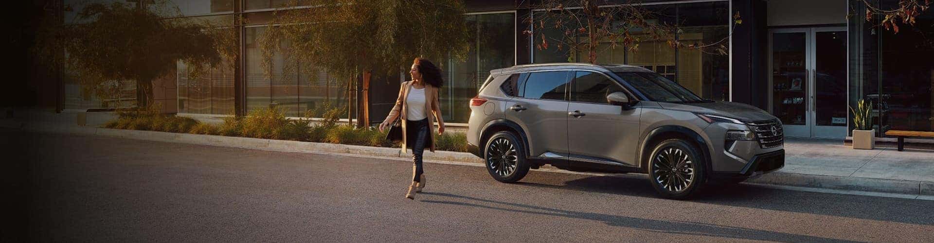 A woman walks confidently on a sunlit street towards a silver SUV parked by a modern building with trees lining the sidewalk.