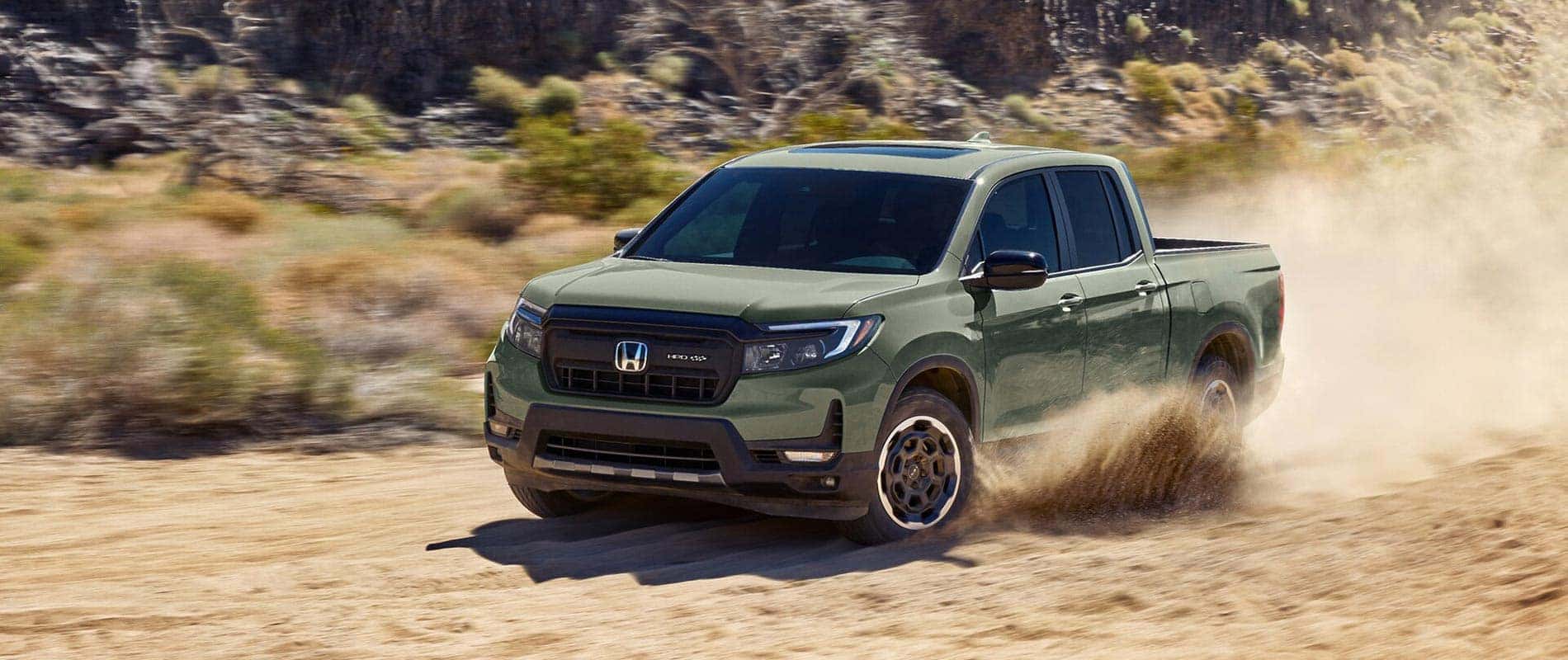 A green 2026 Honda Ridgeline kicking up sand as it drives off-road through the desert.