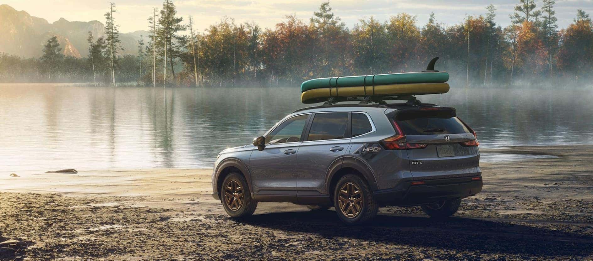 A blue Honda CR-V Hybrid parked on a lake beach with green and yellow surfboards strapped to its roof.