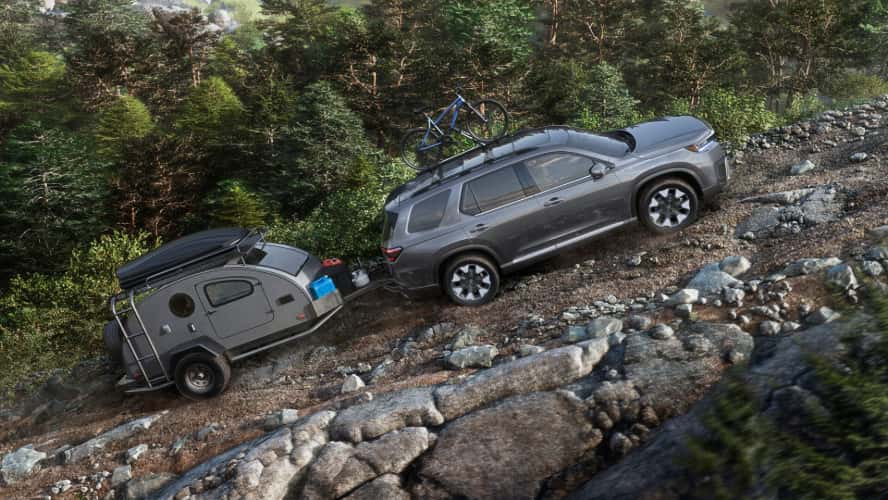 A gray Honda Pilot towing a small trailer uphill on a rocky road, with a blue bike on the roof.