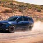 A blue Honda CR-V kicking up dust as it drives on a dirt road through a desert.