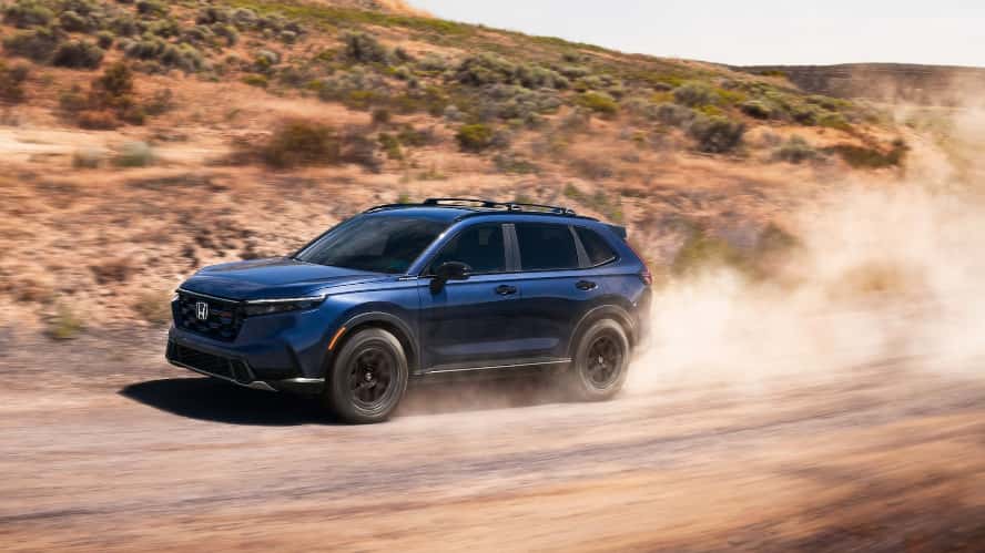 A blue Honda CR-V kicking up dust as it drives on a dirt road through a desert.