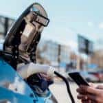 a man kneels next to a charging electric vehicle, smiling