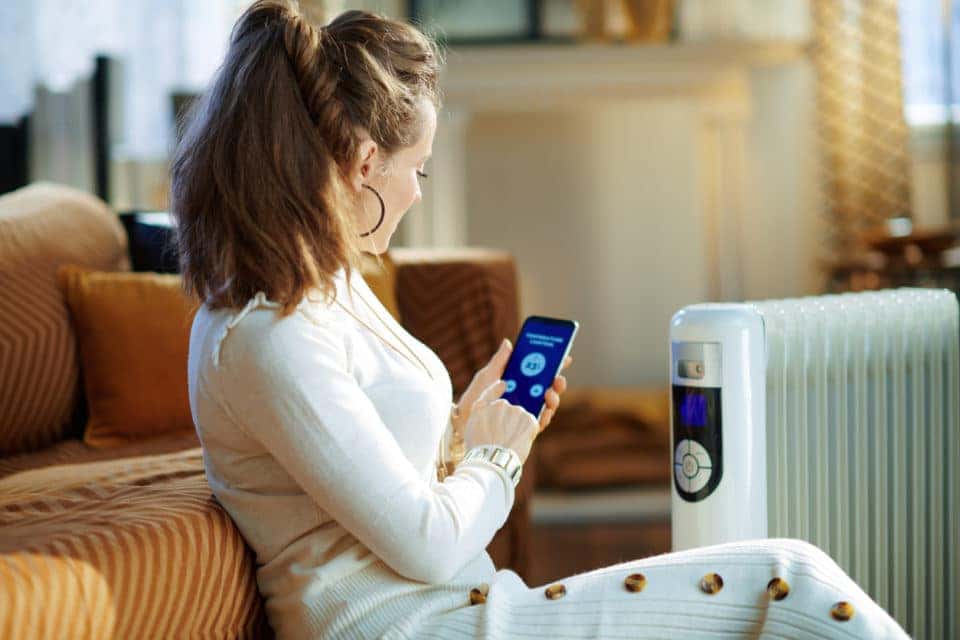 Stylish woman in white sweater and skirt in the modern living room in sunny winter day sitting near couch and white electric oil radiator using smartphone app for temperature control.