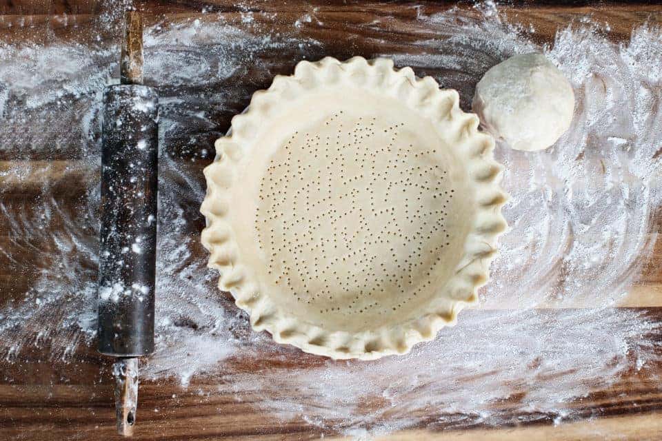 Homemade butter pie crust in pie plate with fluted pinched edge, rolling pin and extra ball of dough over floured rustic wooden background. Crust has been perforated with fork and ready for baking.