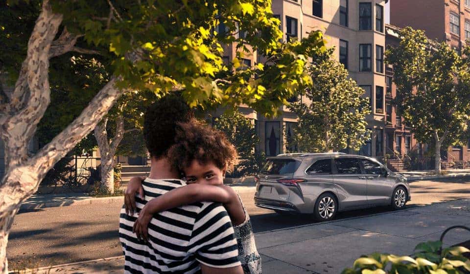 A father and daughter standing on the sidewalk under a tree, their 2021 Toyota Sienna minivan is in the background